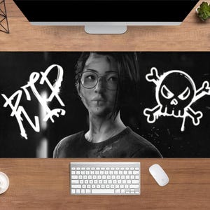 Pode incluir: Tapete de mesa retangular preto e branco com um retrato de uma pessoa usando óculos. O tapete tem o texto "RIP" em estilo grafite e um gráfico de caveira e ossos cruzados. Um teclado e um mouse são visíveis.