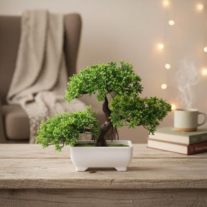 Peut inclure: Un petit bonsaï artificiel avec un feuillage vert vif et un tronc brun est présenté dans un pot rectangulaire blanc. L'arbre est posé sur une surface en bois, avec un arrière-plan flou comprenant une chaise, une couverture et une tasse de café.
