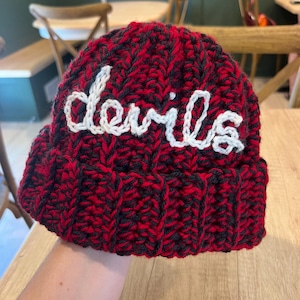May include: A red and black knit beanie with the word "devils" embroidered in white across the front. The hat is made of a thick, textured yarn and has a folded brim. The beanie is a winter accessory.