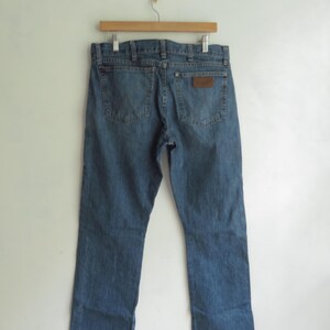 May include: Blue denim jeans hanging on a wooden hanger. The jeans have a classic five-pocket design with a brown leather patch on the back pocket. The jeans are a medium wash with a straight leg fit. The jeans are displayed against a white background.