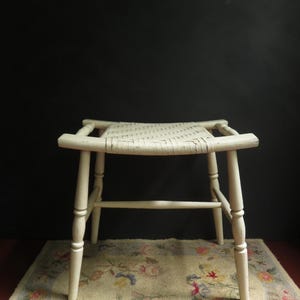 May include: A small, off-white wooden stool featuring a woven seat and four tapered legs connected by horizontal stretchers. The stool has a rectangular frame with slightly extended corners and a smooth, painted finish on the wooden structure.