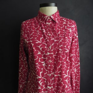 May include: A button-down shirt with a red and white teardrop pattern. The long-sleeved shirt has a single chest pocket and is displayed on a mannequin against a black background.