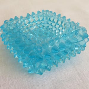 May include: A square, light blue glass ashtray with a textured, spiky exterior. The ashtray has a central depression and is made of transparent glass. The overall design is geometric and decorative.