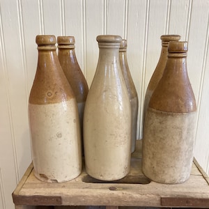 May include: Six vintage stoneware bottles with brown tops in a wooden crate. The bottles are white with brown tops and are arranged in a row.