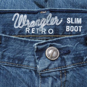 K&ouml;nnte beinhalten: Nahaufnahme einer blauen Jeans. Der Bund zeigt den wei&szlig;en Schriftzug "Wrangler RETRO SLIM BOOT". Ein silberner Knopf mit der Aufschrift "AUTHENTICS" ist sichtbar. Die Jeans hat klassische N&auml;hte und einen Knopfverschluss.