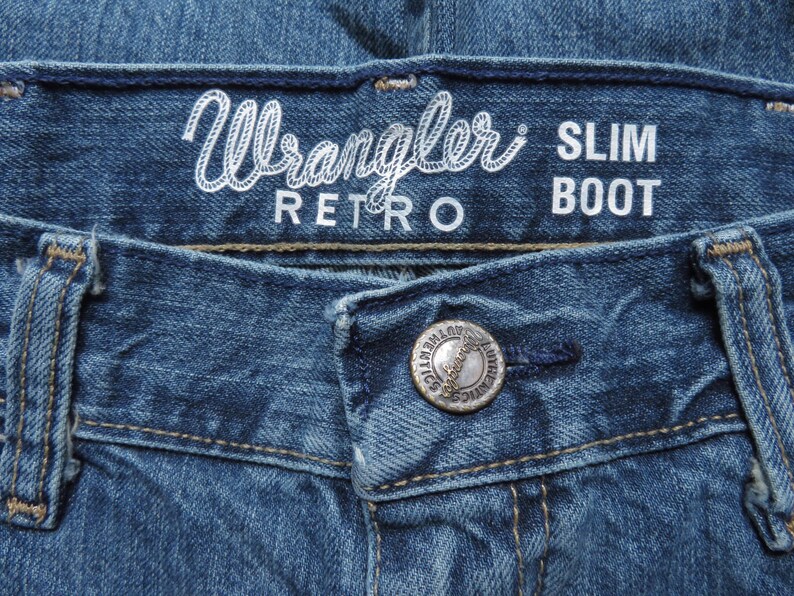 K&ouml;nnte beinhalten: Nahaufnahme einer blauen Jeans. Der Bund zeigt den wei&szlig;en Schriftzug "Wrangler RETRO SLIM BOOT". Ein silberner Knopf mit der Aufschrift "AUTHENTICS" ist sichtbar. Die Jeans hat klassische N&auml;hte und einen Knopfverschluss.