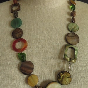 May include: A long necklace with brown, green, and orange round and square beads. The necklace is on a fabric-covered mannequin.