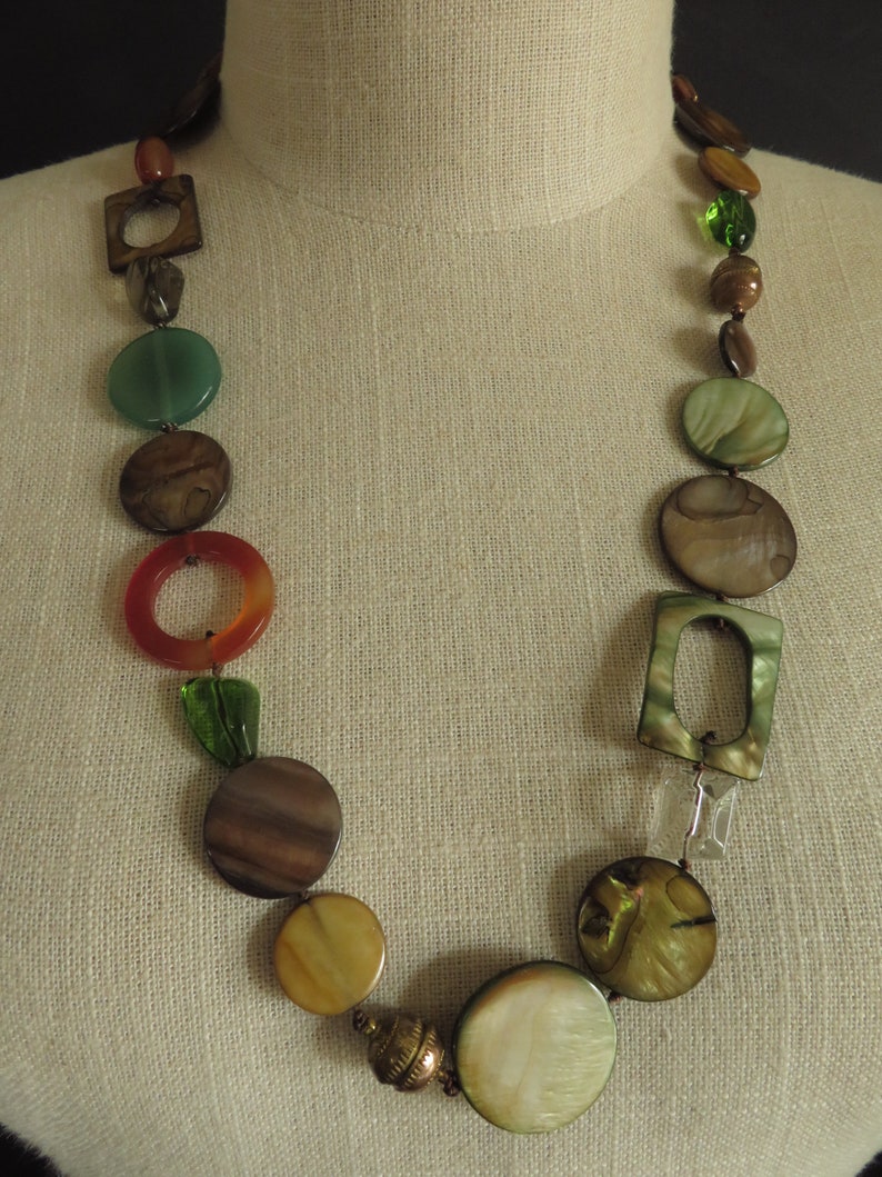 May include: A long necklace with brown, green, and orange round and square beads. The necklace is on a fabric-covered mannequin.