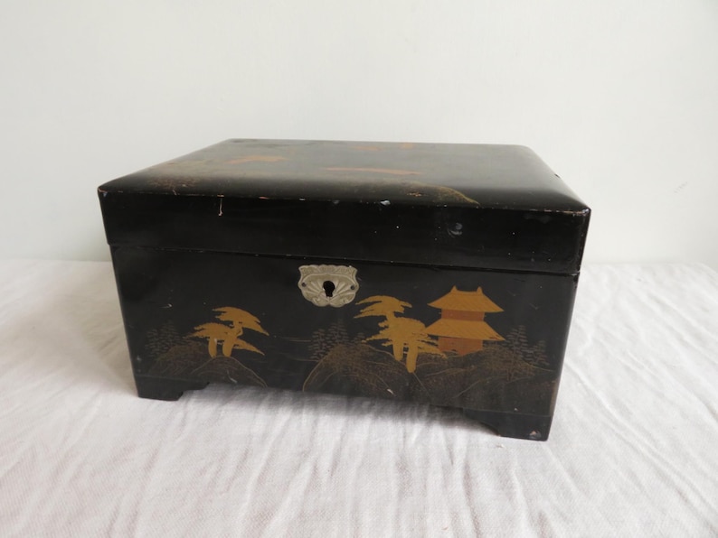 K&ouml;nnte beinhalten: Eine schwarze Holzkiste mit einem gold-schwarzen Design eines japanischen Landschaftsbildes. Die Kiste hat ein silbernes Schloss und ein Schl&uuml;sselloch.