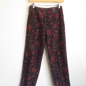 May include: Dark navy floral print pants with red and olive green accents. The pants are hanging on a wooden hanger against a white background. The pants have a button closure and a straight leg design.