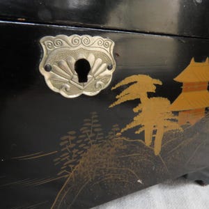 K&ouml;nnte beinhalten: Schwarze Holzkiste mit einem silbernen Metallverschluss und einem japanisch inspirierten Design. Die Kiste zeigt goldfarbene B&auml;ume und eine Pagode.