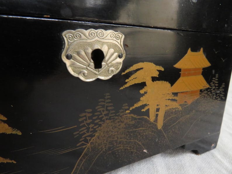 K&ouml;nnte beinhalten: Schwarze Holzkiste mit einem silbernen Metallverschluss und einem japanisch inspirierten Design. Die Kiste zeigt goldfarbene B&auml;ume und eine Pagode.