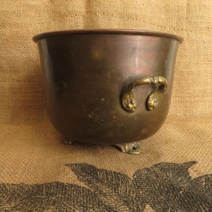 May include: A vintage copper planter with a rounded body, ornate handles, and small feet. The planter has a dark, aged patina, and the handles feature a decorative design. The background is a textured, tan fabric.