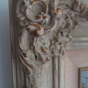 Puede incluir: Primer plano de una esquina de un marco de fotos vintage. El marco est&aacute; pintado en un color blanquecino desgastado con detalles dorados. Las tallas y detalles ornamentados son visibles, mostrando una est&eacute;tica cl&aacute;sica y antigua.
