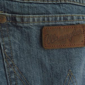 K&ouml;nnte beinhalten: Nahaufnahme von blauen Jeans aus Denim mit einem braunen Lederaufn&auml;her. Der Aufn&auml;her zeigt den Markennamen "Wrangler" in Schreibschrift. Sichtbare N&auml;hte und ein Metallknopf an der Ges&auml;&szlig;tasche.