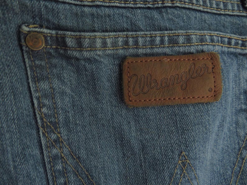 K&ouml;nnte beinhalten: Nahaufnahme von blauen Jeans aus Denim mit einem braunen Lederaufn&auml;her. Der Aufn&auml;her zeigt den Markennamen "Wrangler" in Schreibschrift. Sichtbare N&auml;hte und ein Metallknopf an der Ges&auml;&szlig;tasche.