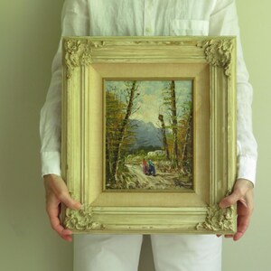Puede incluir: Una pintura de paisaje enmarcada, de estilo antiguo. La obra representa un camino a trav&eacute;s de un bosque con figuras a lo lejos. El marco es ornamentado, con un acabado crema y detalles decorativos en las esquinas. La pintura mide unos 30 cm por 25 cm.