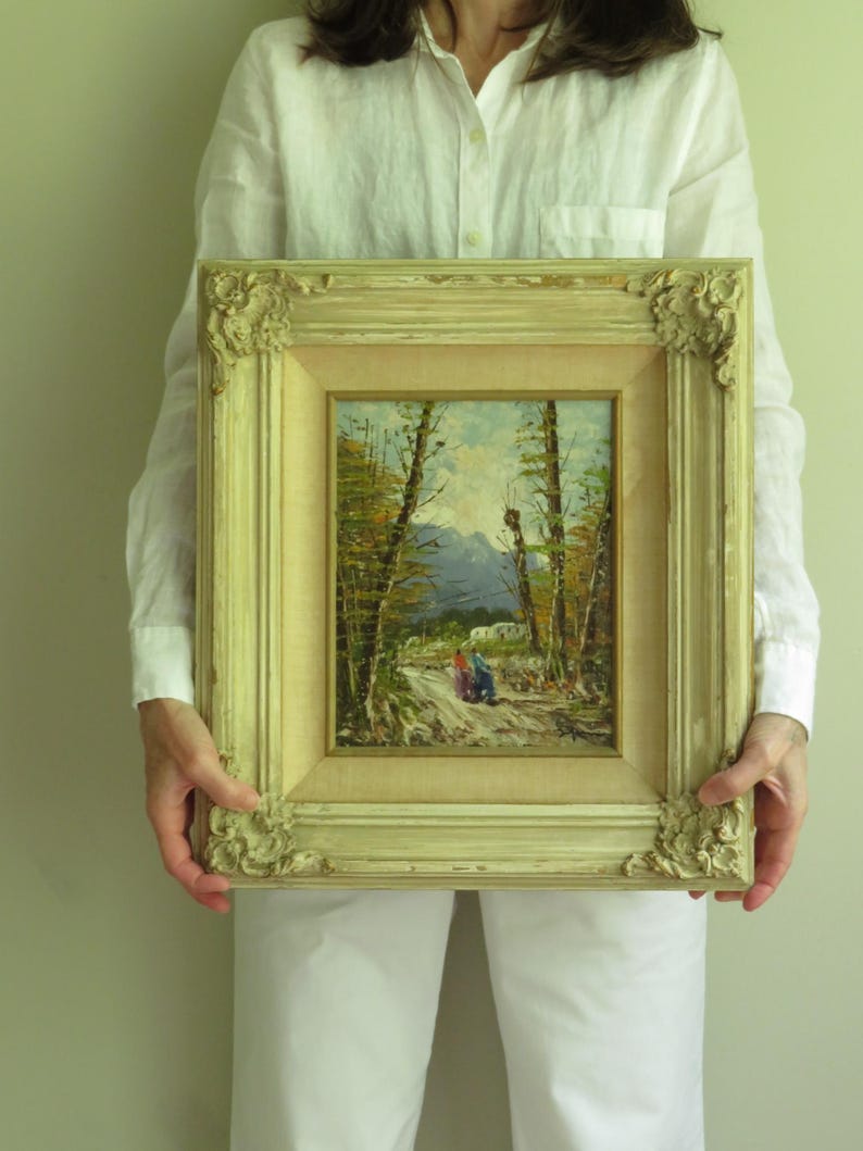 Puede incluir: Una pintura de paisaje enmarcada, de estilo antiguo. La obra representa un camino a trav&eacute;s de un bosque con figuras a lo lejos. El marco es ornamentado, con un acabado crema y detalles decorativos en las esquinas. La pintura mide unos 30 cm por 25 cm.