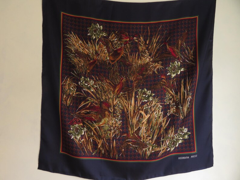 Puede incluir: Un pa&ntilde;uelo cuadrado de seda con un borde azul marino y un dise&ntilde;o central con p&aacute;jaros, plantas y flores en tonos marrones, verdes y rojos. El pa&ntilde;uelo tiene un fondo estampado y el texto "HERMANA PUCCI" en la esquina inferior derecha.