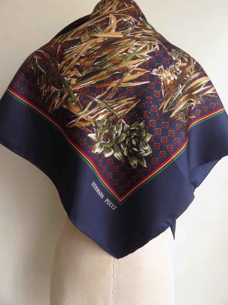 Puede incluir: Un pa&ntilde;uelo cuadrado de seda con borde azul marino y un estampado floral y bot&aacute;nico en tonos marr&oacute;n, verde y beige. El pa&ntilde;uelo tiene un ribete rojo y amarillo y el texto "HERMANA PUCCI" en blanco.