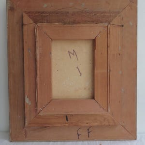 Puede incluir: Parte trasera de una obra de arte enmarcada con un marco de madera marr&oacute;n claro. La obra de arte est&aacute; asegurada con grapas. La parte trasera de la obra de arte tiene las letras "M" e "I" escritas con un rotulador azul. El marco tiene las letras "FF" escritas con rotulador negro.