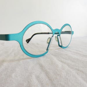 May include: Teal-colored eyeglasses with a unique, round frame design. The glasses have clear lenses and black temples. The frame is a vibrant teal, and the glasses are set against a white background.