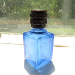 May include: A small, square-shaped cobalt blue glass bottle featuring a faceted design on its sides. The bottle is topped with a cylindrical, dark metallic cap that has a textured, knurled band around its center, providing a distinct contrast to the smooth, vibrant blue glass body.