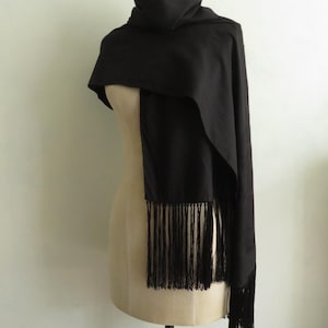 May include: A solid black rectangular scarf draped over a mannequin. The scarf features a smooth, matte texture and is finished with long, thin, matching black fringe detailing along the bottom edges, creating a clean and uniform appearance.