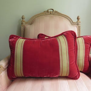 May include: Two rectangular red velvet pillows with a central vertical stripe of olive green and gold fabric. The pillows are on a pink upholstered chair with a carved wooden back.