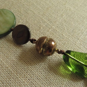 May include: A close-up of a necklace with green, brown, and gold beads. The beads are strung on a brown cord. The necklace is lying on a beige fabric surface.