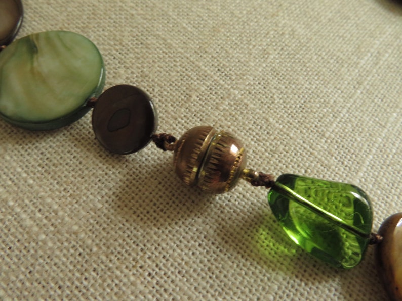 May include: A close-up of a necklace with green, brown, and gold beads. The beads are strung on a brown cord. The necklace is lying on a beige fabric surface.