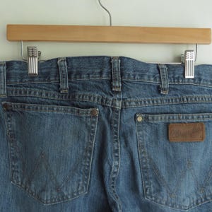 K&ouml;nnte beinhalten: Blaue Jeans aus Denim mit zwei Ges&auml;&szlig;taschen und einem braunen Leder-Wrangler-Patch. Die Jeans h&auml;ngen an einem Holzb&uuml;gel mit Metallclips.