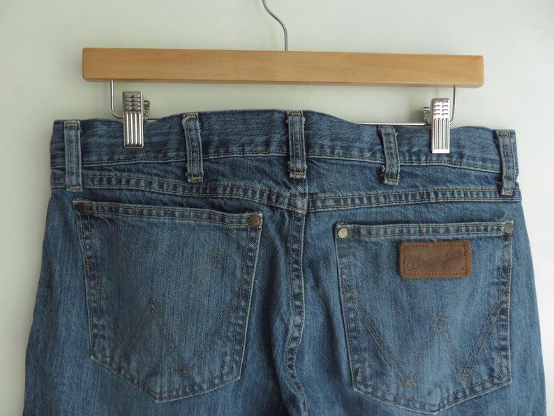 K&ouml;nnte beinhalten: Blaue Jeans aus Denim mit zwei Ges&auml;&szlig;taschen und einem braunen Leder-Wrangler-Patch. Die Jeans h&auml;ngen an einem Holzb&uuml;gel mit Metallclips.