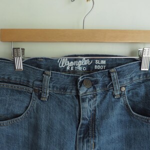 K&ouml;nnte beinhalten: Nahaufnahme einer blauen Wrangler Retro Slim Boot Jeans, die an einem Holzb&uuml;gel h&auml;ngt. Die Jeans hat ein klassisches F&uuml;nf-Taschen-Design, einen Knopfverschluss und das Wrangler-Logo &uuml;ber der Vordertasche.