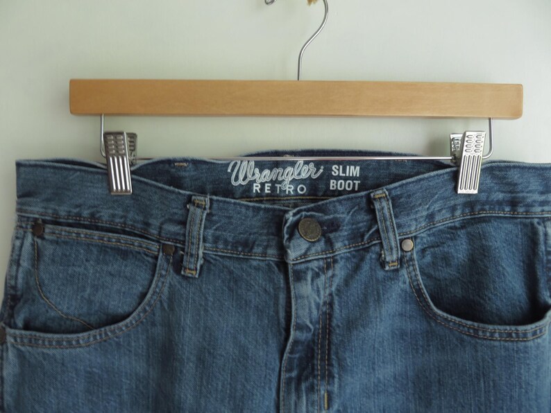 K&ouml;nnte beinhalten: Nahaufnahme einer blauen Wrangler Retro Slim Boot Jeans, die an einem Holzb&uuml;gel h&auml;ngt. Die Jeans hat ein klassisches F&uuml;nf-Taschen-Design, einen Knopfverschluss und das Wrangler-Logo &uuml;ber der Vordertasche.