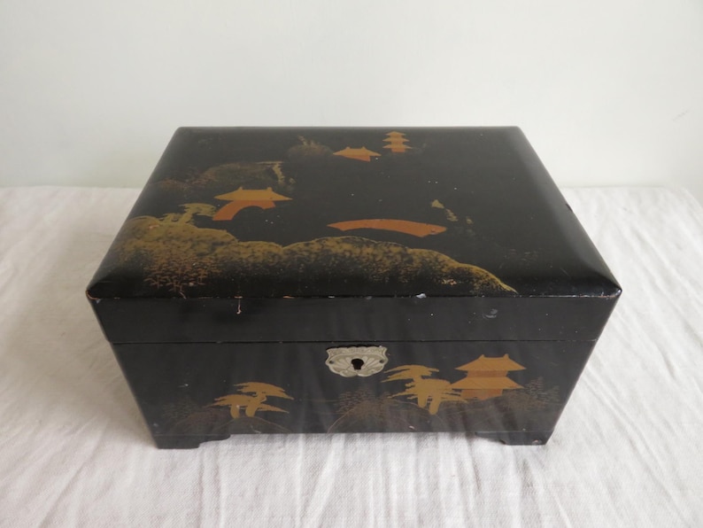 K&ouml;nnte beinhalten: Schwarze Holzschatulle mit einem goldenen und orangefarbenen japanischen Landschaftsdesign auf dem Deckel. Die Schatulle hat ein kleines Schloss und ein Scharnier aus Metall.