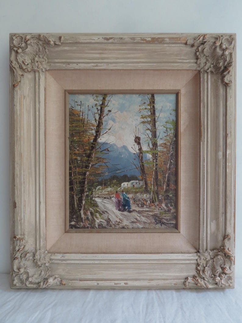 Puede incluir: Pintura al &oacute;leo de un paisaje con un camino, &aacute;rboles y monta&ntilde;as. La obra de arte est&aacute; enmarcada en un marco desgastado, ornamentado, blanquecino. La pintura representa dos figuras caminando por un sendero.