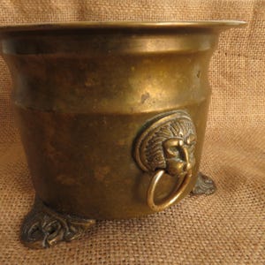 May include: A vintage brass planter with a wide rim and a lion's head ring handle. The planter has three decorative feet and a weathered finish, suggesting age and use. The overall shape is cylindrical.