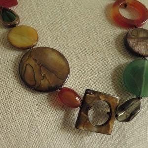 May include: A multi-colored necklace with round and square shaped beads. The beads are made of different materials, including wood, stone, and shell. The necklace is brown, green, red, and yellow.