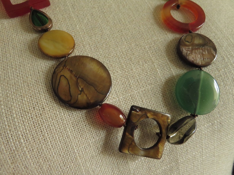 May include: A multi-colored necklace with round and square shaped beads. The beads are made of different materials, including wood, stone, and shell. The necklace is brown, green, red, and yellow.