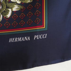 Puede incluir: Pa&ntilde;uelo de seda azul marino con un dise&ntilde;o floral en tonos verdes, marrones y blancos. El pa&ntilde;uelo presenta un borde geom&eacute;trico con detalles en rojo y verde. El texto "HERMANA PUCCI" est&aacute; impreso en blanco.