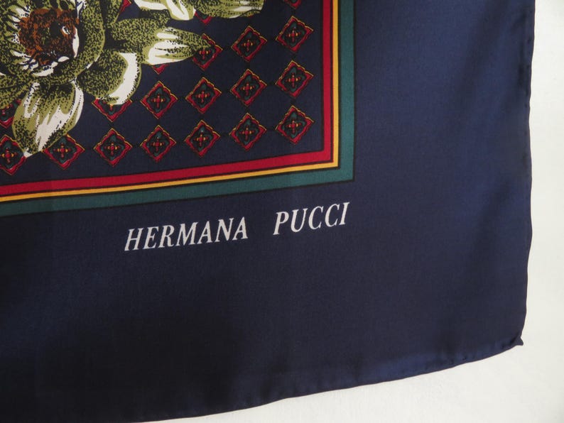 Puede incluir: Pa&ntilde;uelo de seda azul marino con un dise&ntilde;o floral en tonos verdes, marrones y blancos. El pa&ntilde;uelo presenta un borde geom&eacute;trico con detalles en rojo y verde. El texto "HERMANA PUCCI" est&aacute; impreso en blanco.