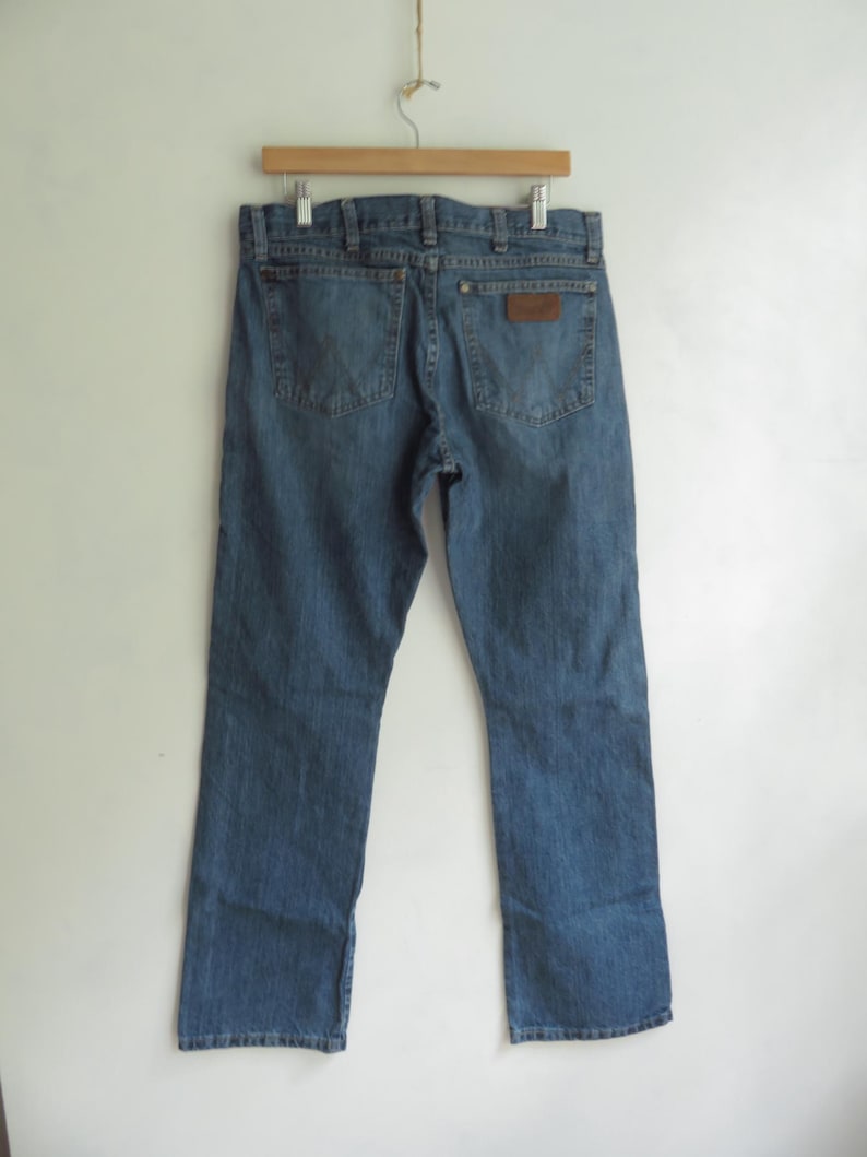 May include: Blue denim jeans hanging on a wooden hanger. The jeans have a classic five-pocket design with a brown leather patch on the back pocket. The jeans are a medium wash with a straight leg fit. The jeans are displayed against a white background.