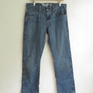 K&ouml;nnte beinhalten: Blaue Wrangler Retro Slim Boot Jeans aus Denim, die an einem Holzb&uuml;gel h&auml;ngen. Die Jeans haben ein klassisches F&uuml;nf-Taschen-Design und eine verwaschene Optik. Das Wrangler-Logo ist auf der Vordertasche zu sehen.