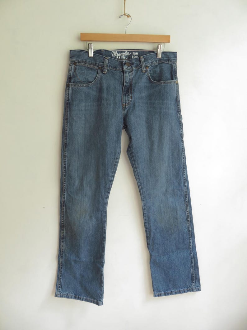 K&ouml;nnte beinhalten: Blaue Wrangler Retro Slim Boot Jeans aus Denim, die an einem Holzb&uuml;gel h&auml;ngen. Die Jeans haben ein klassisches F&uuml;nf-Taschen-Design und eine verwaschene Optik. Das Wrangler-Logo ist auf der Vordertasche zu sehen.