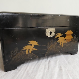 K&ouml;nnte beinhalten: Eine schwarze Holzkiste mit einem gold-schwarzen Design aus B&auml;umen und einer Pagode. Die Kiste hat einen silbernen Metallverschluss.