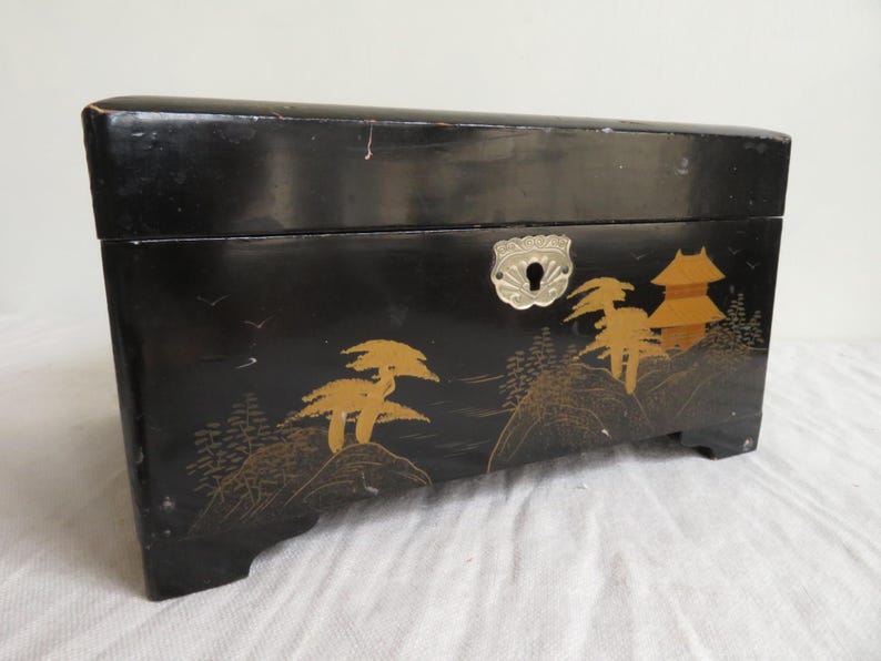 K&ouml;nnte beinhalten: Eine schwarze Holzkiste mit einem gold-schwarzen Design aus B&auml;umen und einer Pagode. Die Kiste hat einen silbernen Metallverschluss.