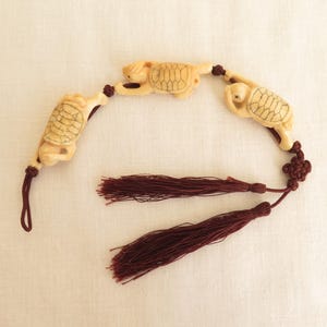 May include: Ivory-colored turtle charm with a dark red tassel. The turtles are carved with detailed shell patterns and connected by a dark red cord. The tassel is made of multiple strands of dark red thread.