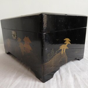 K&ouml;nnte beinhalten: Eine schwarze Holzkiste mit einem gold-schwarzen Design aus B&auml;umen und einem Berg. Die Kiste hat einen Metallverschluss und Scharniere.