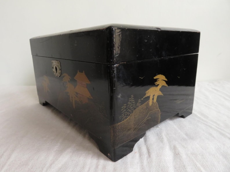 K&ouml;nnte beinhalten: Eine schwarze Holzkiste mit einem gold-schwarzen Design aus B&auml;umen und einem Berg. Die Kiste hat einen Metallverschluss und Scharniere.